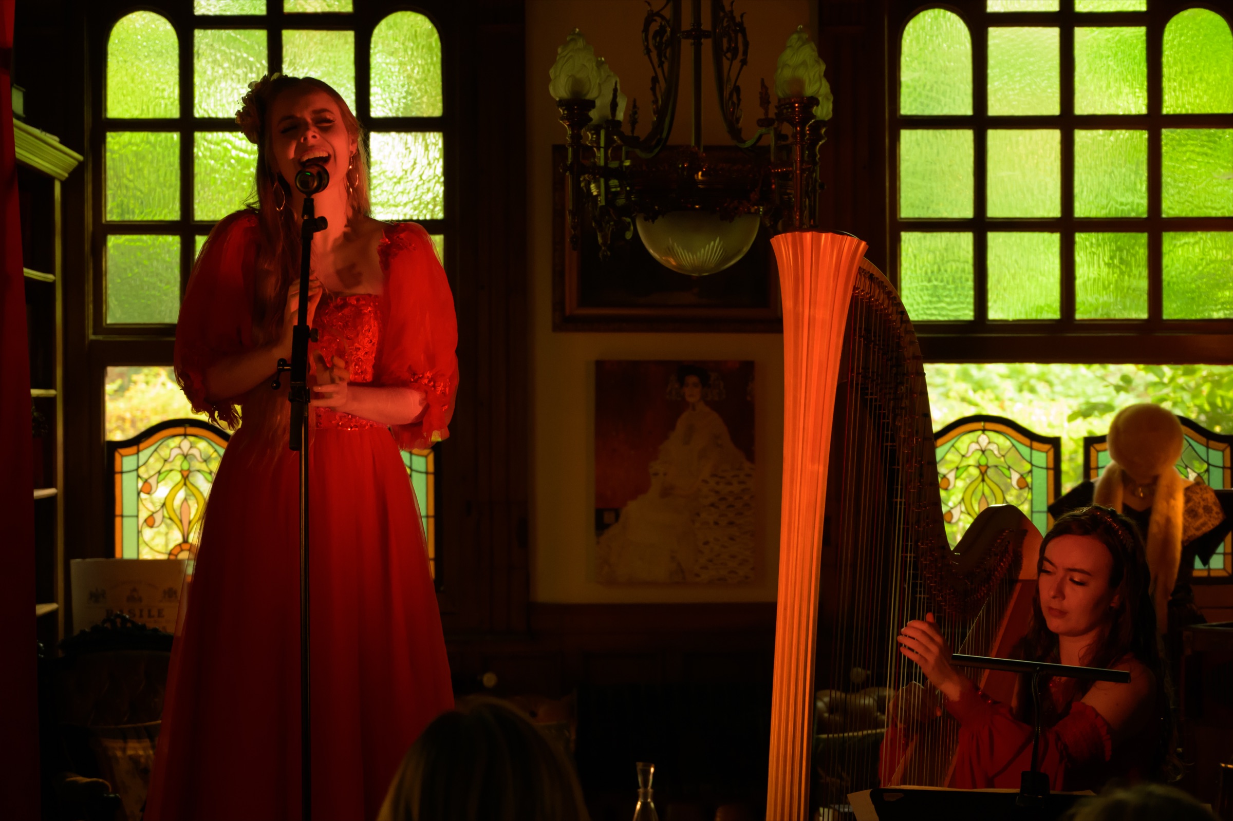 Singer in a red gown at the microphone and a harpist, in a hall with green stained-glass windows at Villa Madonna