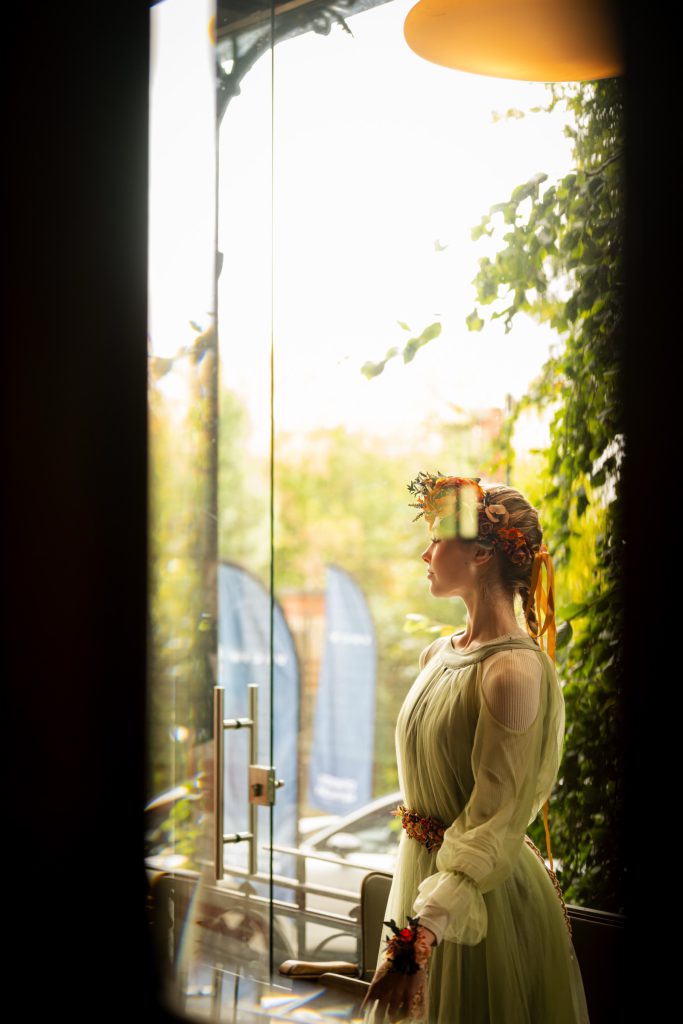 Aurelia in a sage gown and flower headpiece by a sunlit glass door