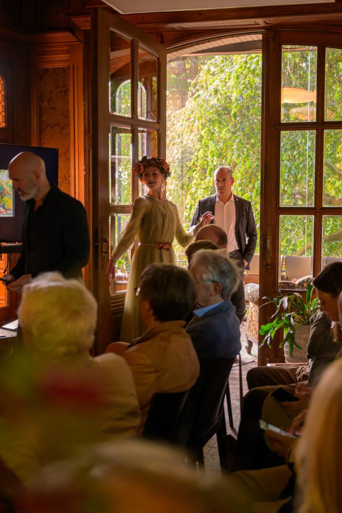 Aurelia leading guests past the paintings into the main hall, garden light behind her