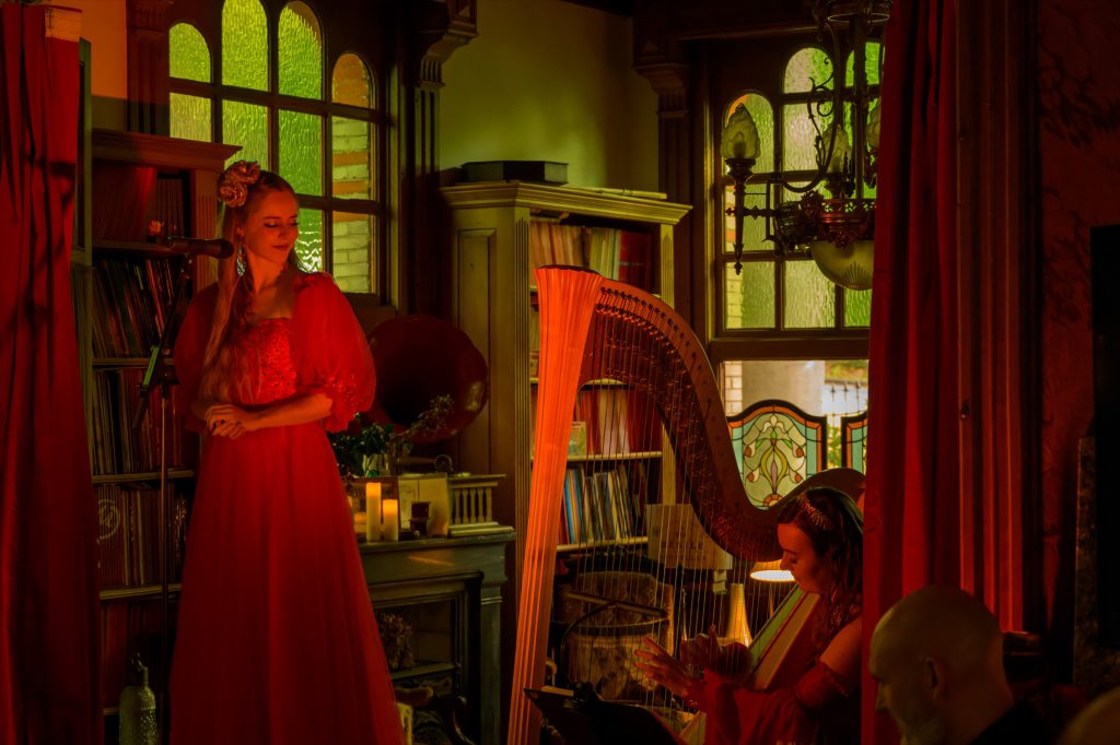Singer in red and harpist performing between red curtains and bookshelves