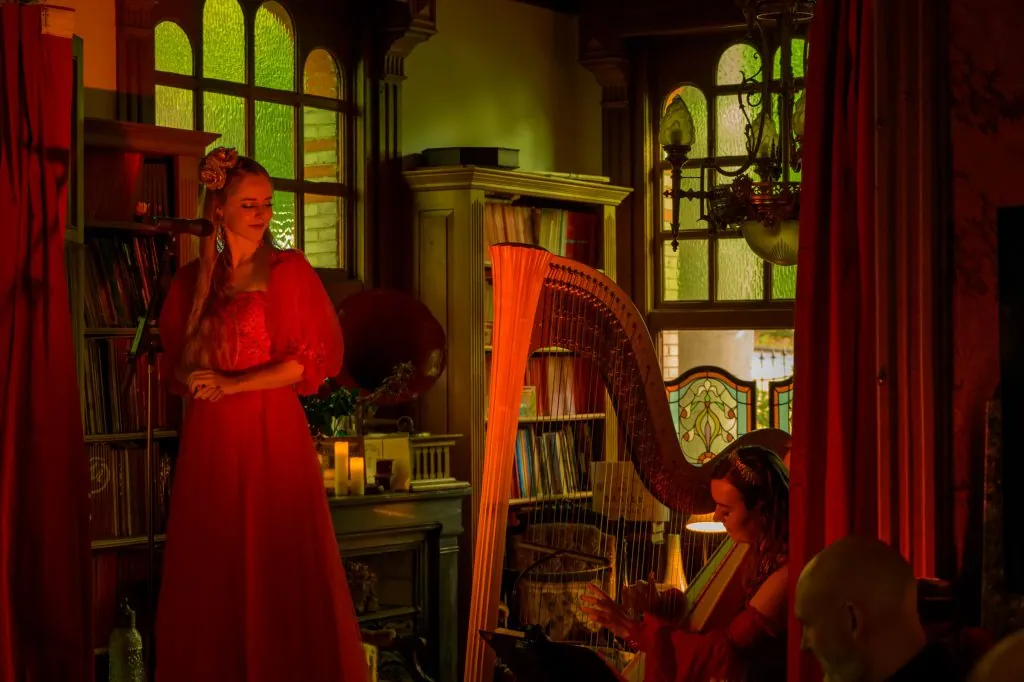 Singer in red and harpist performing between red curtains and bookshelves