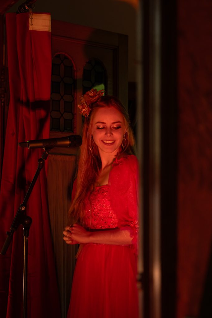 The singer in a red gown smiling beside the microphone, lit in warm red light