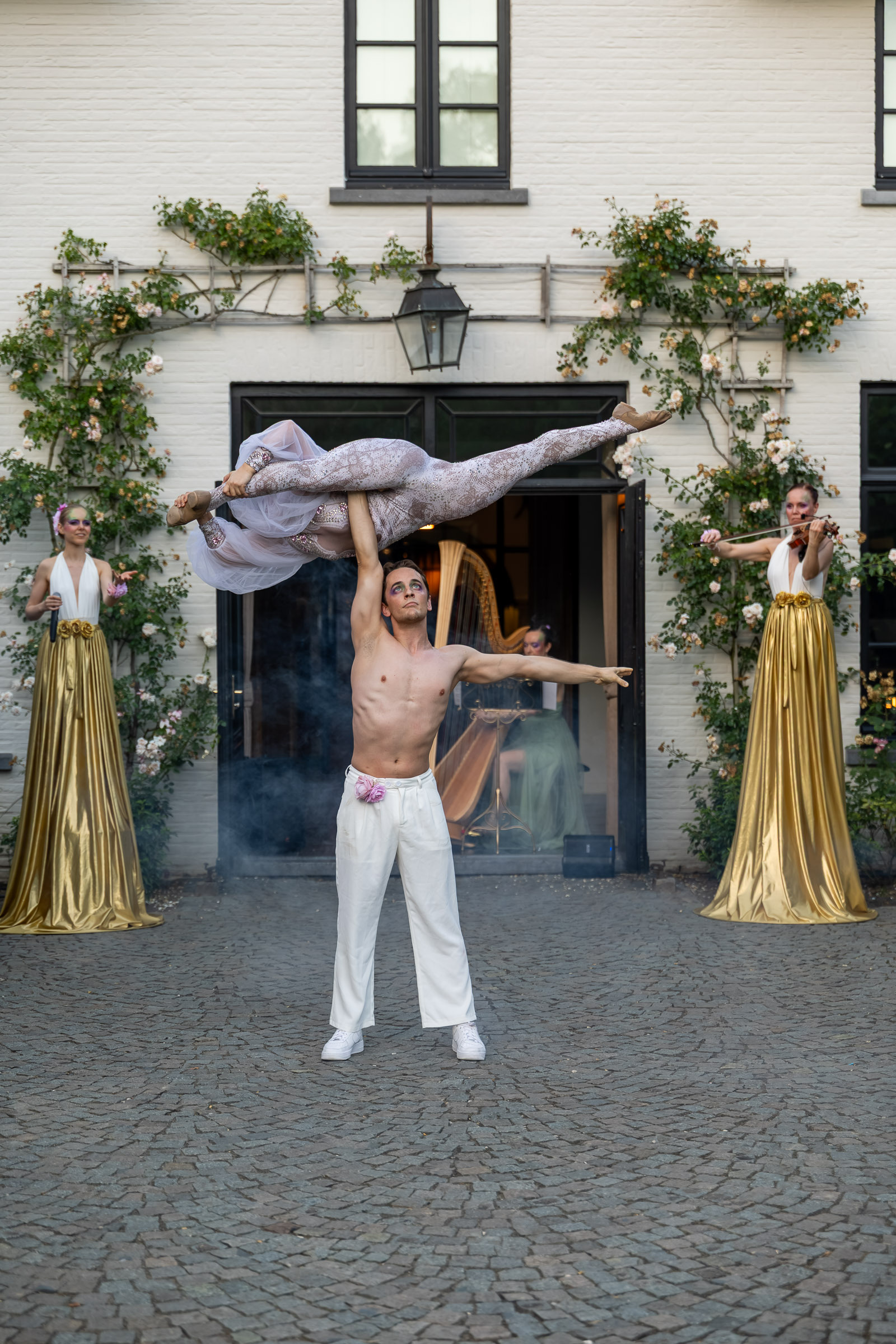 An acrobat lifting a partner overhead in the courtyard of a villa, flanked by two stilt walkers in gold