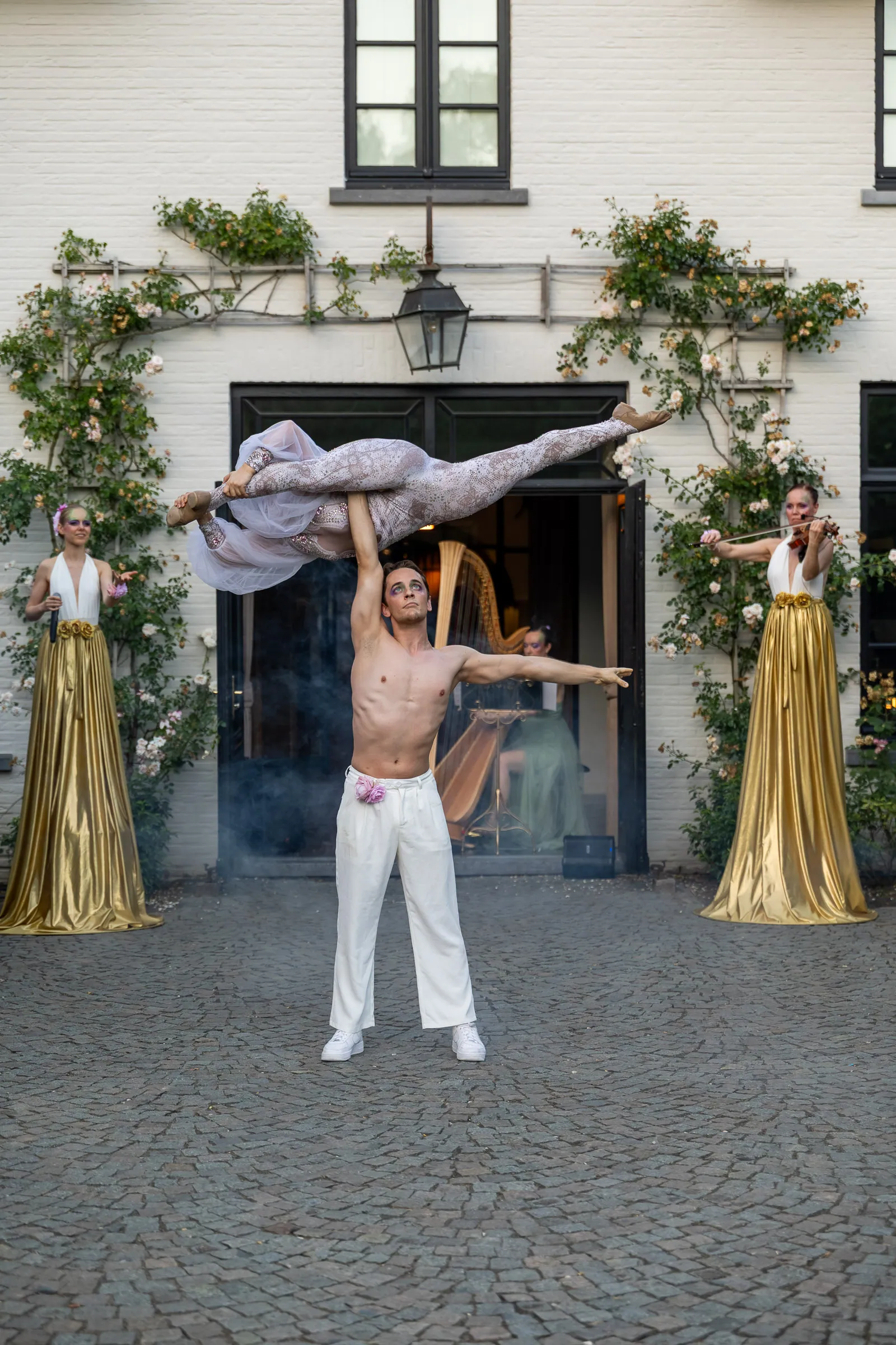 An acrobat lifting a partner overhead in the courtyard of a villa, flanked by two stilt walkers in gold