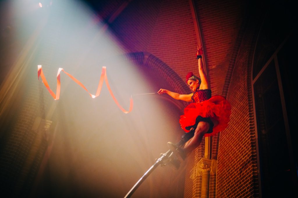 An aerial acrobat in red climbing silks in a beam of light against a brick wall
