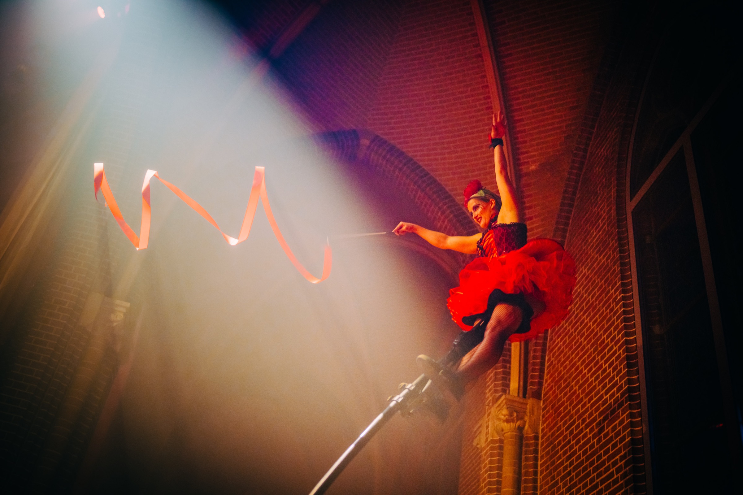 An aerial acrobat in red climbing silks in a beam of light against a brick wall