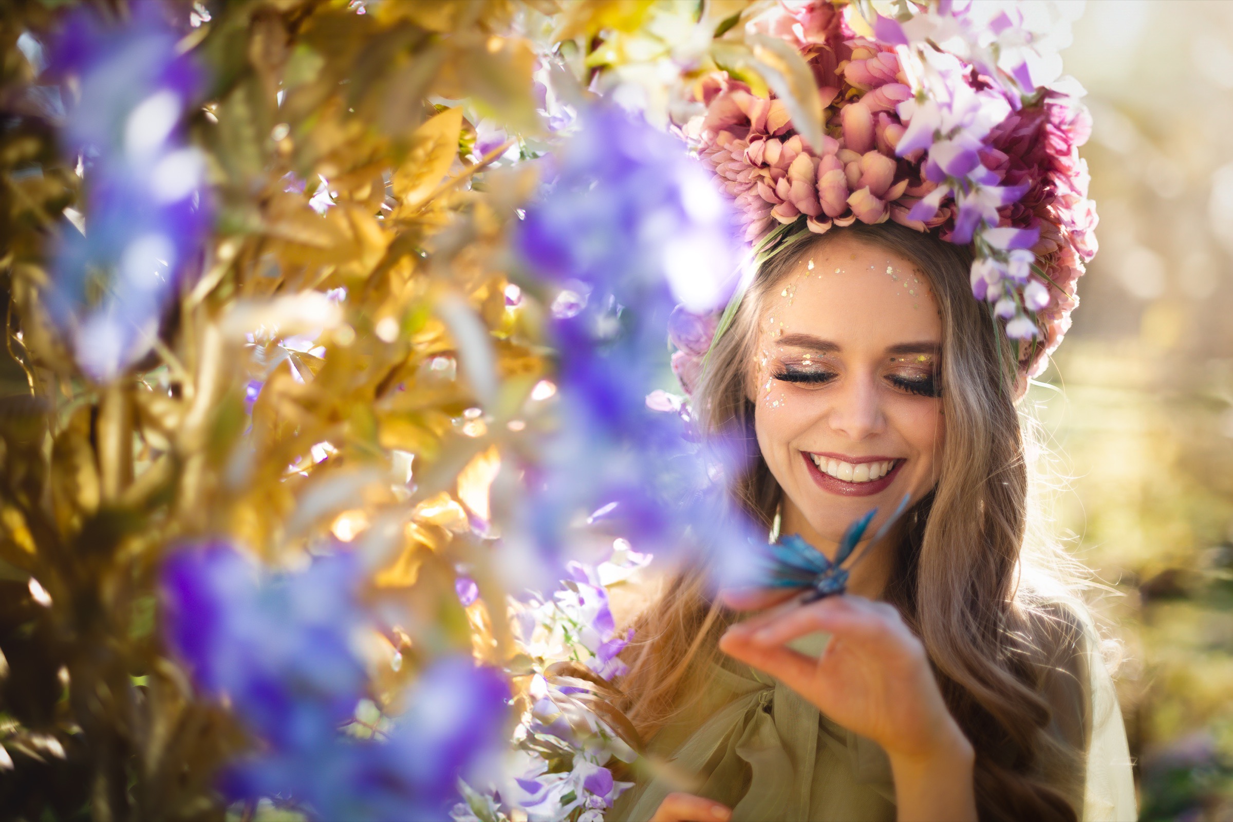 A performer in a crown of pink flowers smiling among golden leaves and purple blossoms
