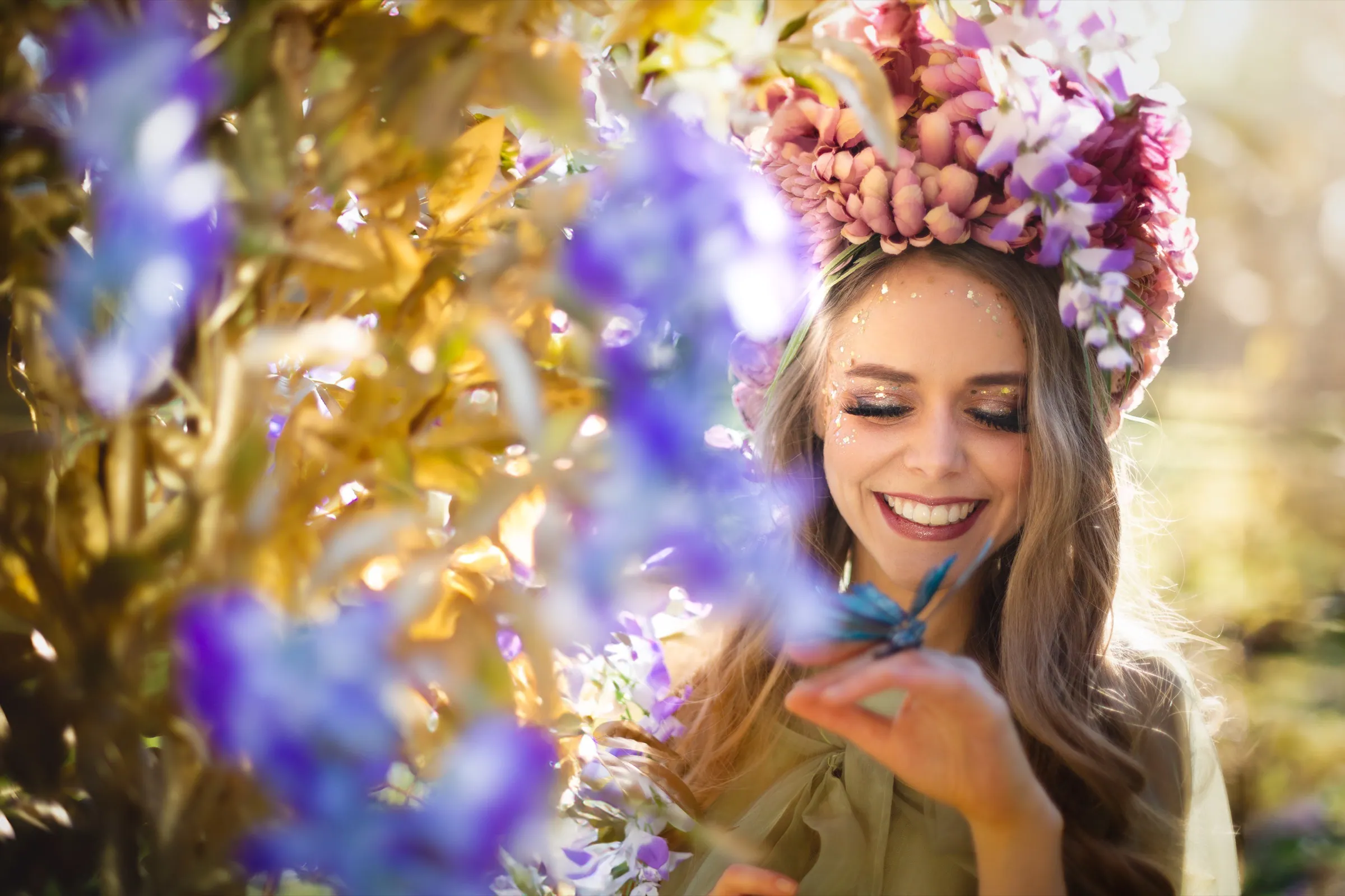 A performer in a crown of pink flowers smiling among golden leaves and purple blossoms