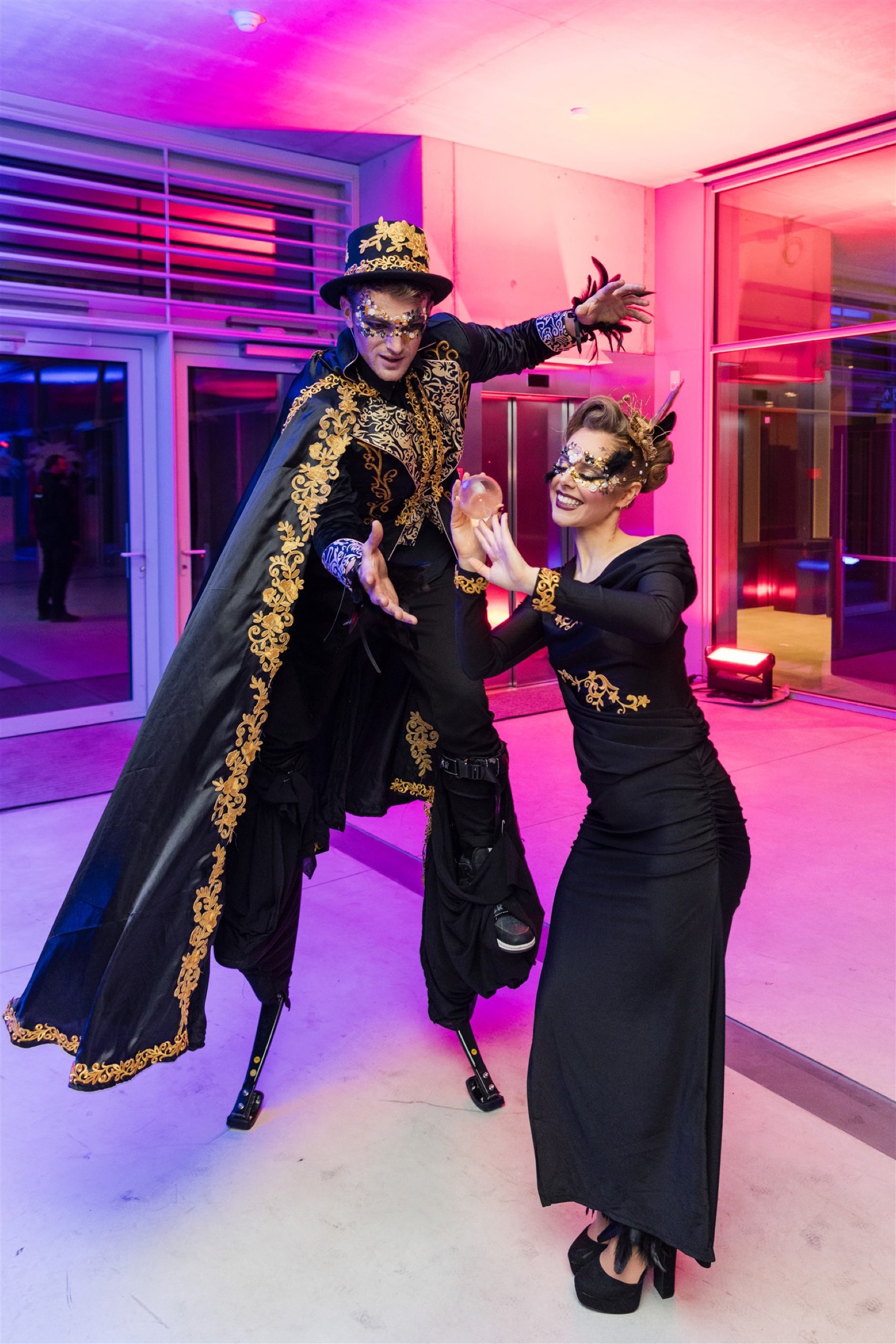 A stiltwalker in a black and gold cape posing with a masked performer in black, in pink light