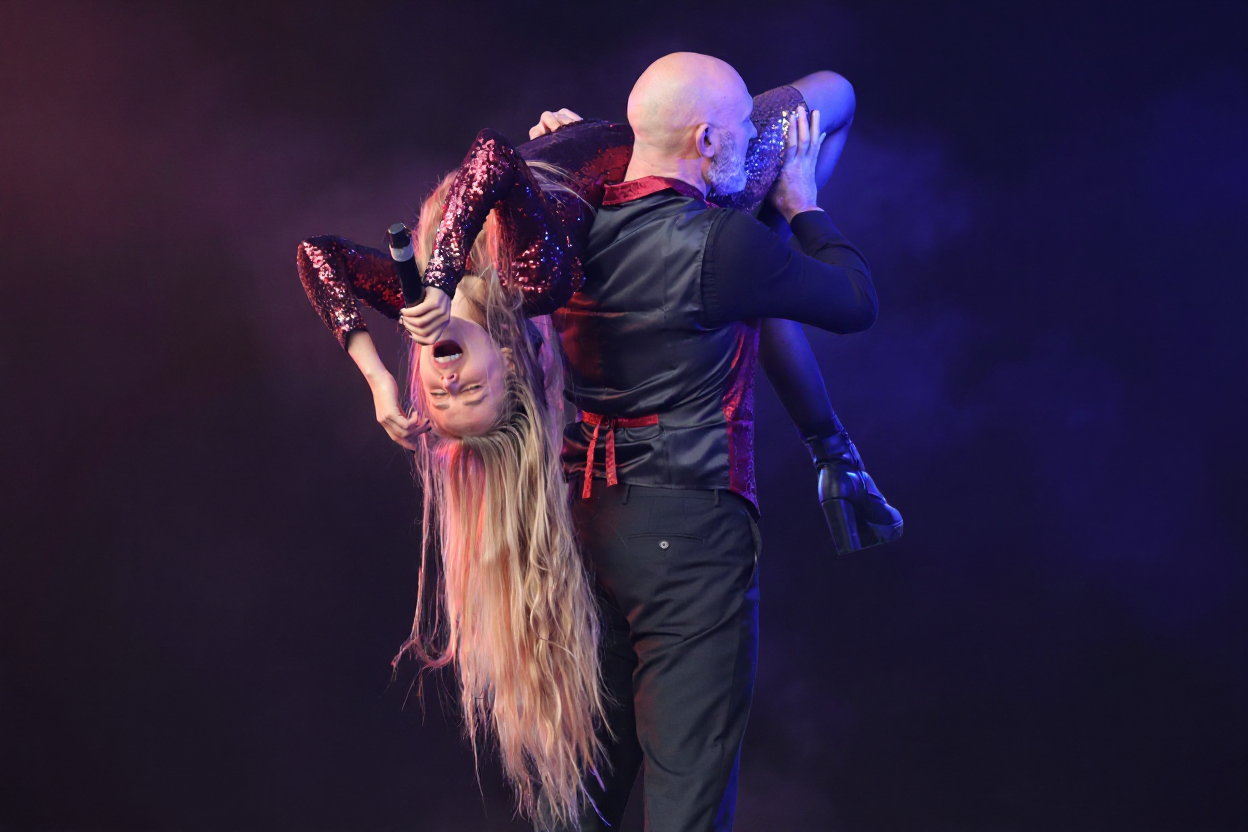 A singer in a long gown carried over the shoulder of a performer in a suit, in blue stage light