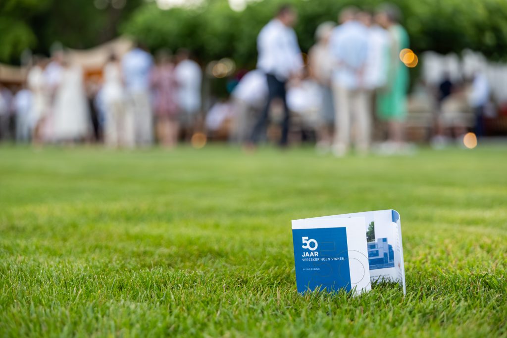 A Verzekeringen Vinken 50-year card on the lawn, guests gathering in the garden behind it