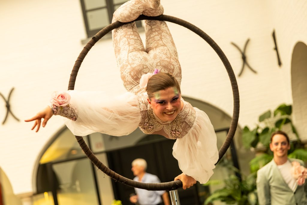 A lyra acrobat in a white costume hanging in her ring, smiling at the guests below