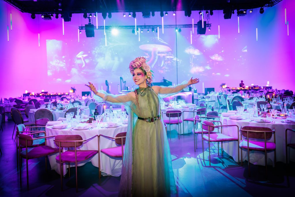 Aurelia in a sage gown with arms wide in a banquet hall lit in pink and blue