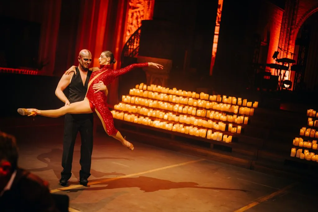 A dancer in a red sequinned dress lifted by her partner in front of a wall of candles