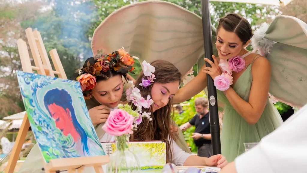 Two elves in flower crowns at a painting easel in the garden, one with large pale wings