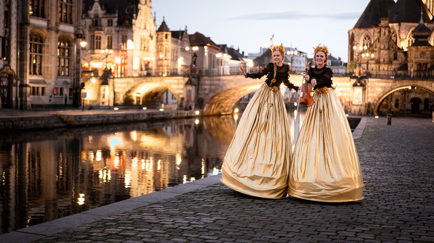 Two stilt performers in gold gowns walking a canal-side street at dusk