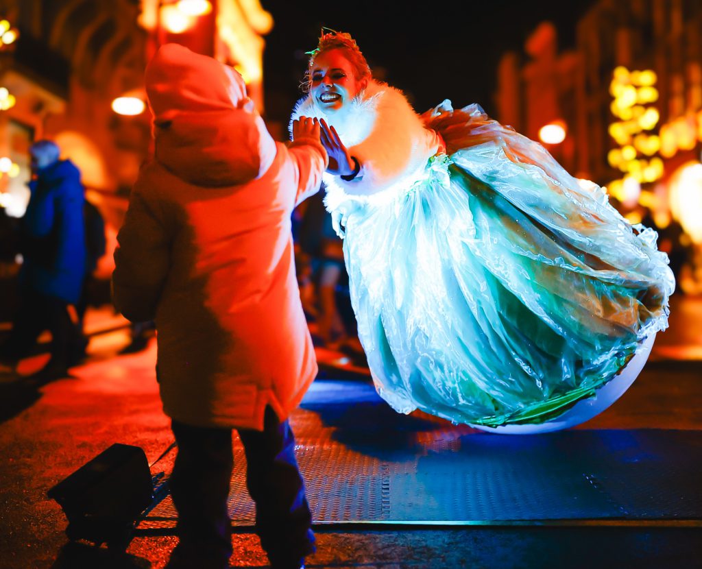 A swayball performer in a glowing blue gown giving a child a high five at a night market