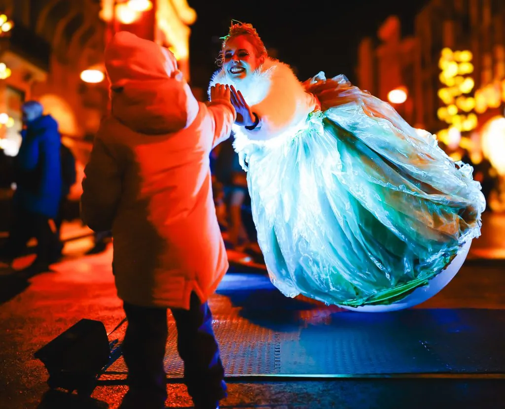 A swayball performer in a glowing blue gown giving a child a high five at a night market