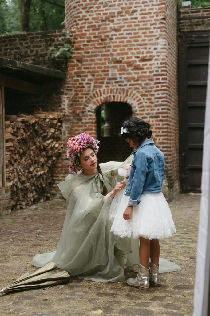 A performer in a sage green gown and flower crown kneeling to talk to a young girl in a courtyard