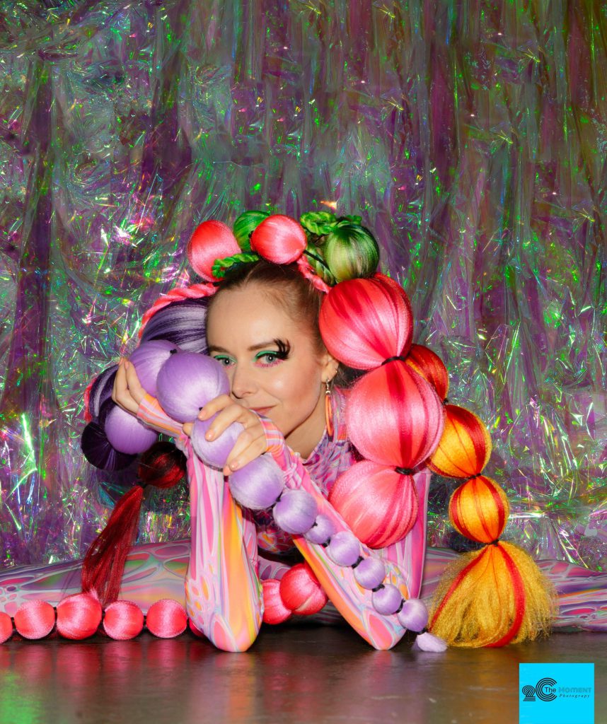 A performer in a wig of colourful balloons and a lilac costume, posing against an iridescent backdrop