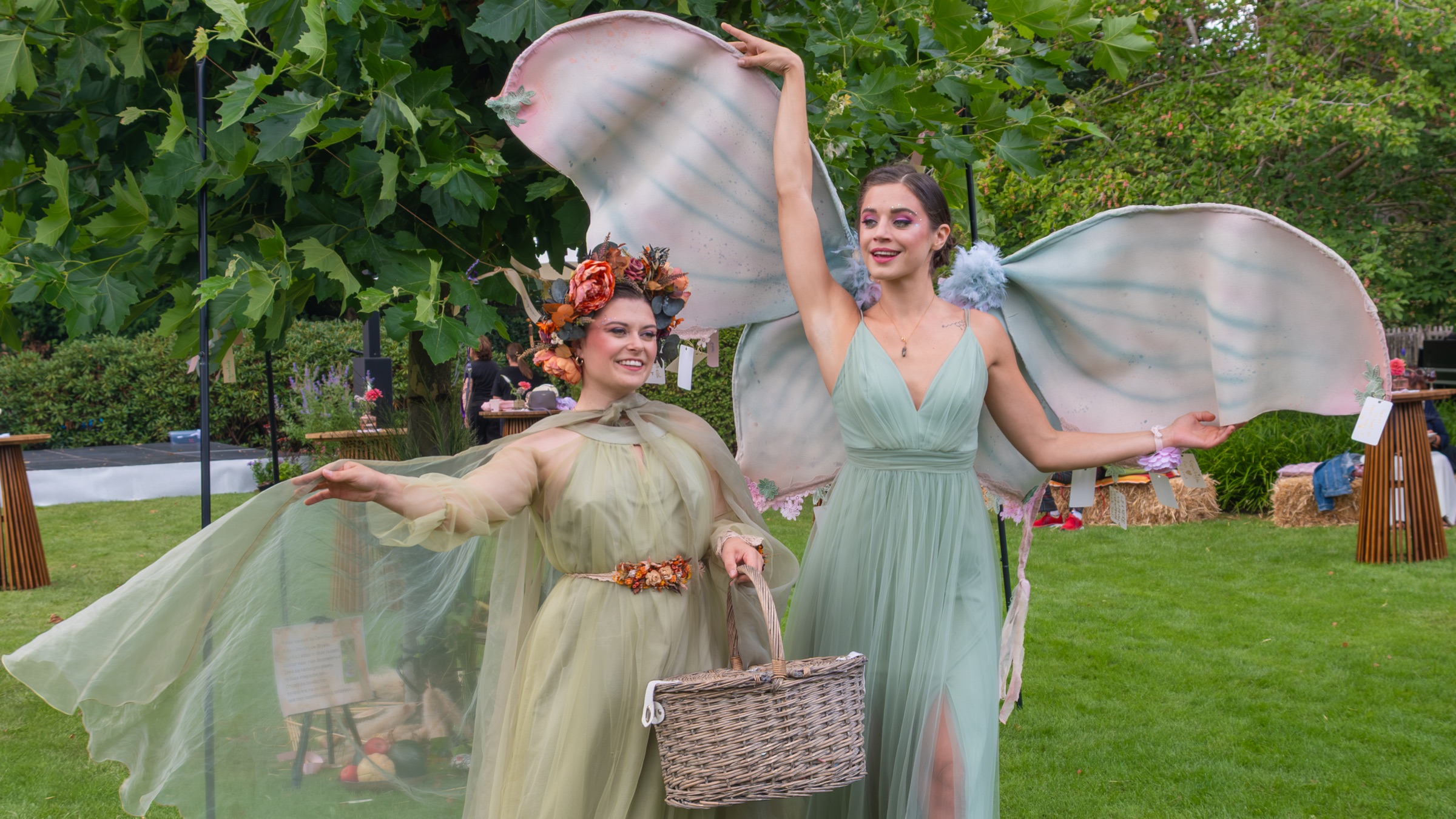 Two performers in green dresses with translucent wings walking across a lawn