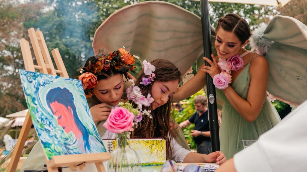 Two performers in flower crowns and pastel wings painting a portrait at an easel at an outdoor event