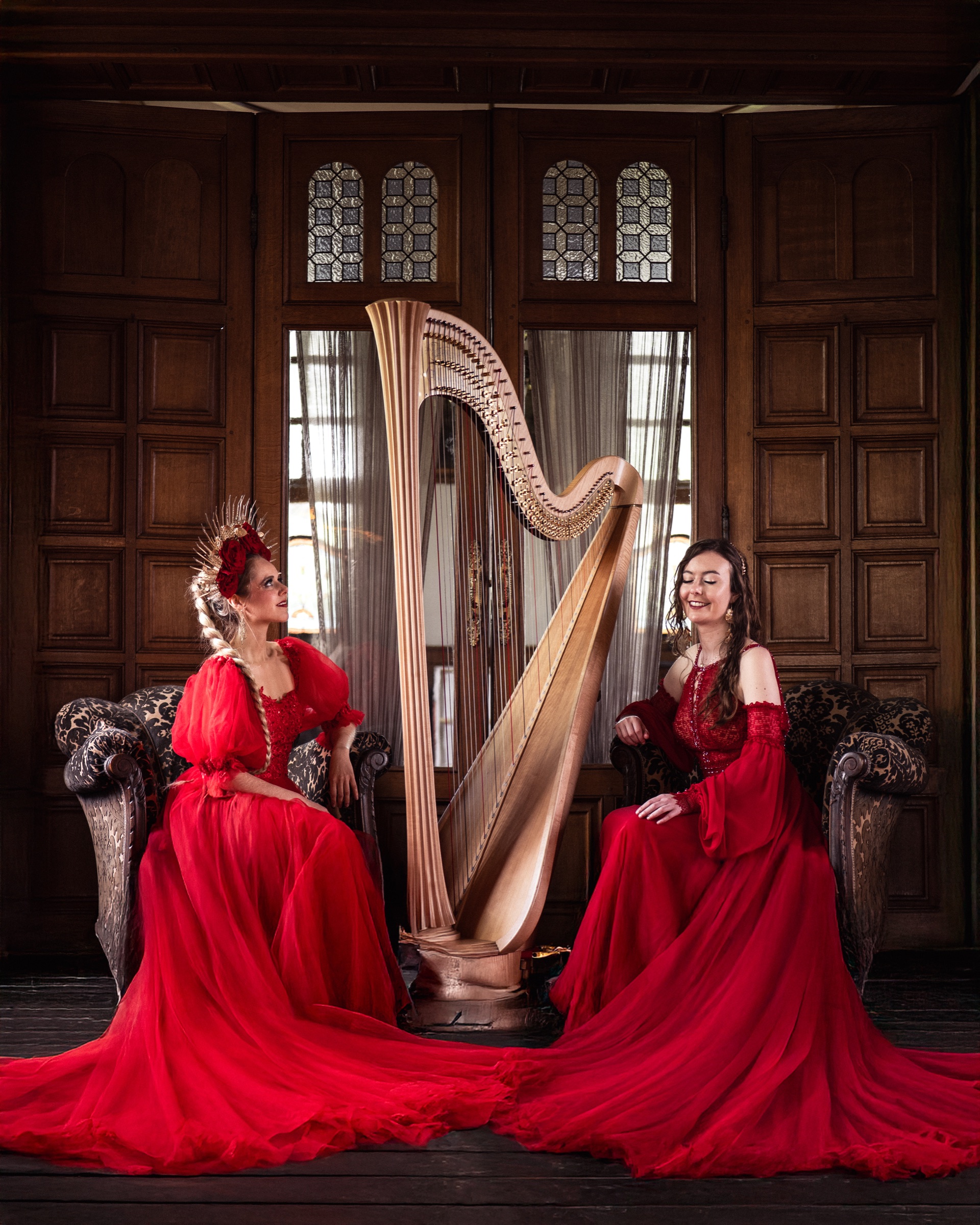 A singer and a harpist in red gowns beside a golden harp in a wood-panelled room