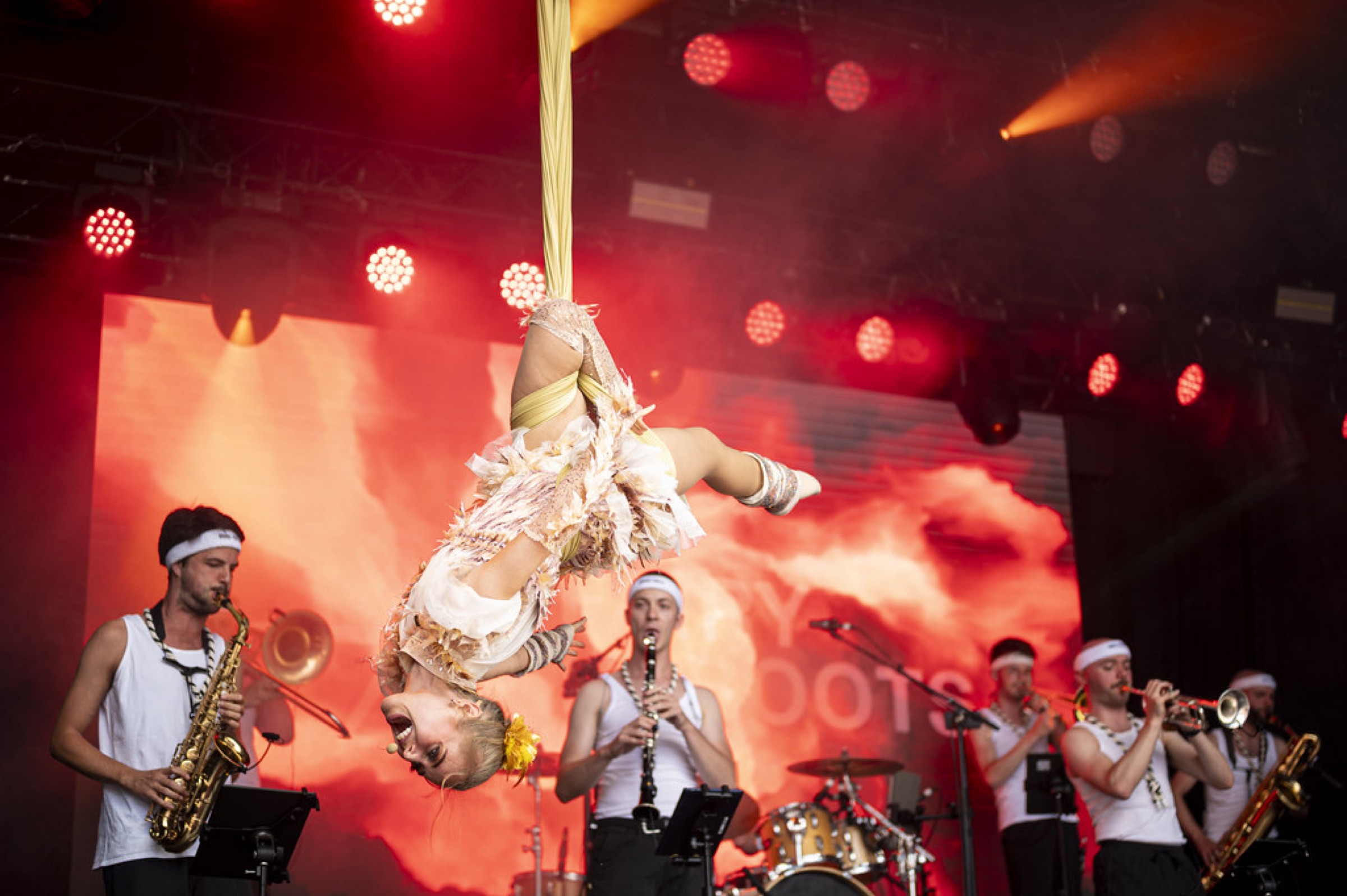 An aerial acrobat hanging upside down above a brass band on a red-lit festival stage