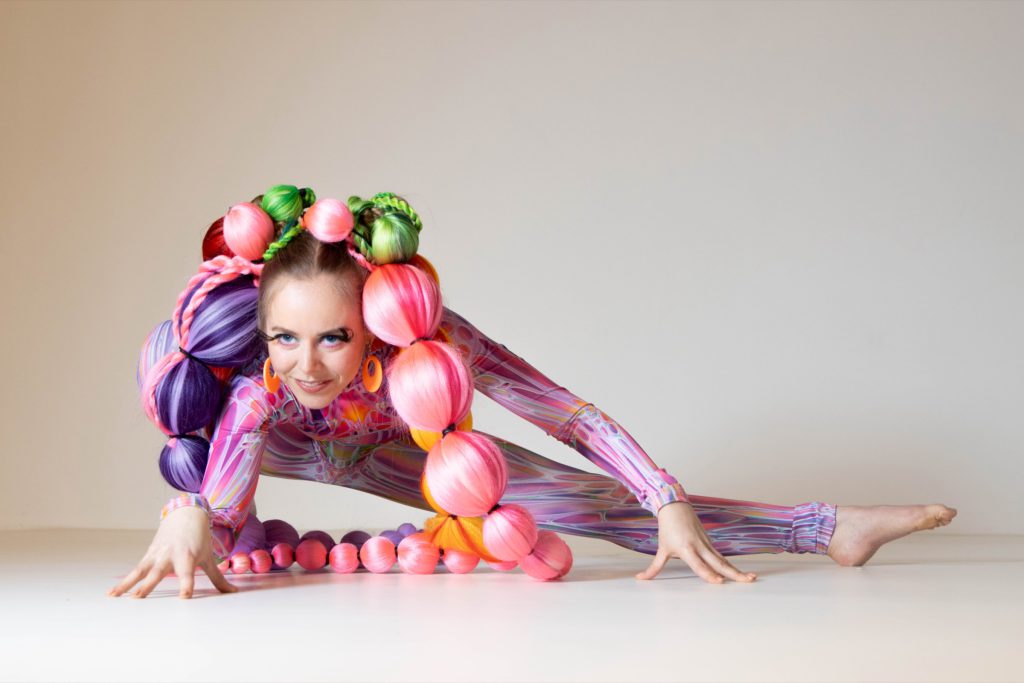 A contortionist in a pink and violet bodysuit and balloon costume, stretched on a white floor